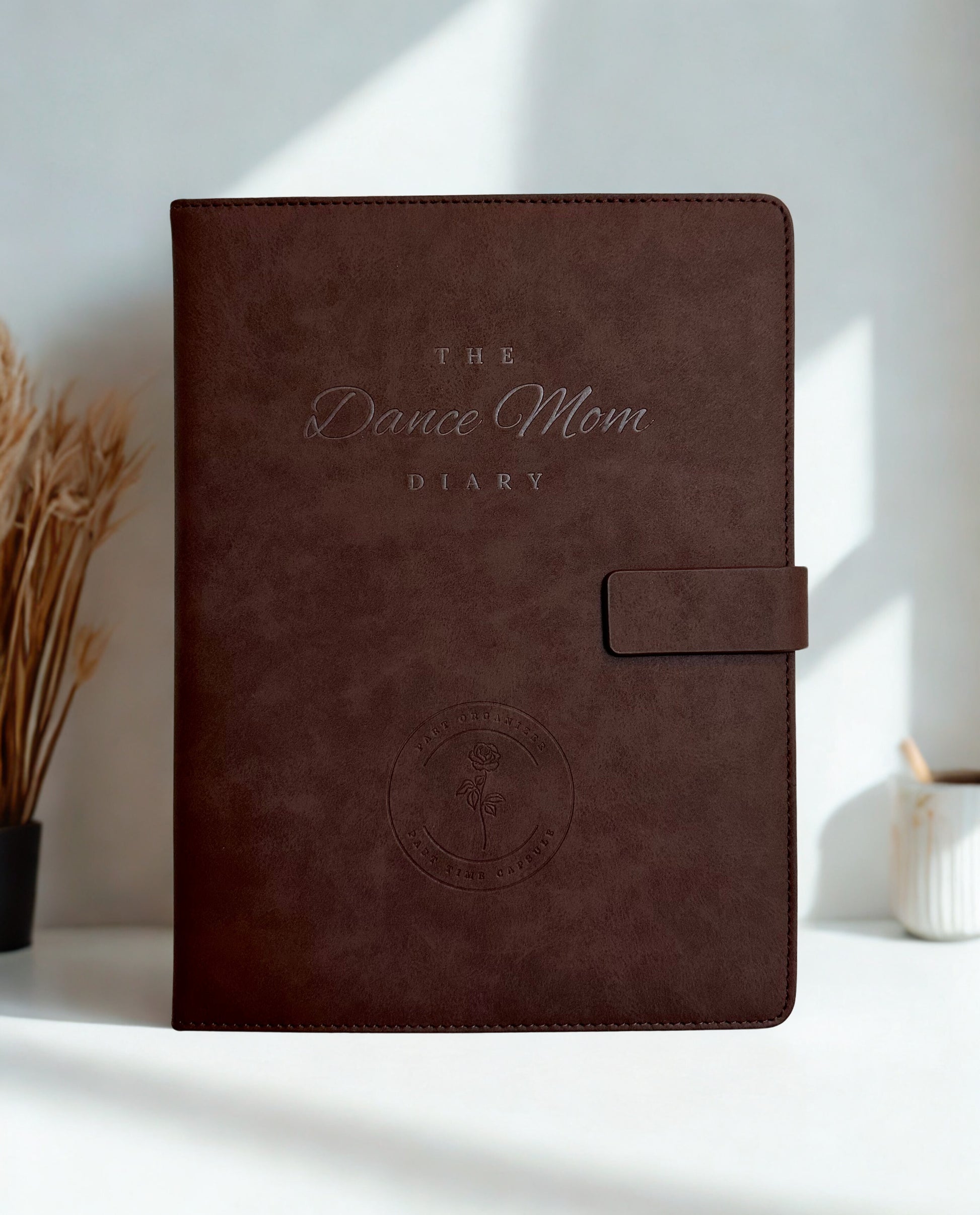 Brown leather Dance Mom Diary with embossed cover and strap closure with a white background and natural lighting.