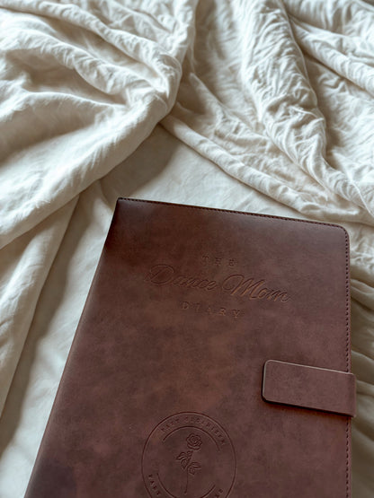 Brown leather Dance Mom Diary with embossed cover and strap closure on a white textured surface.