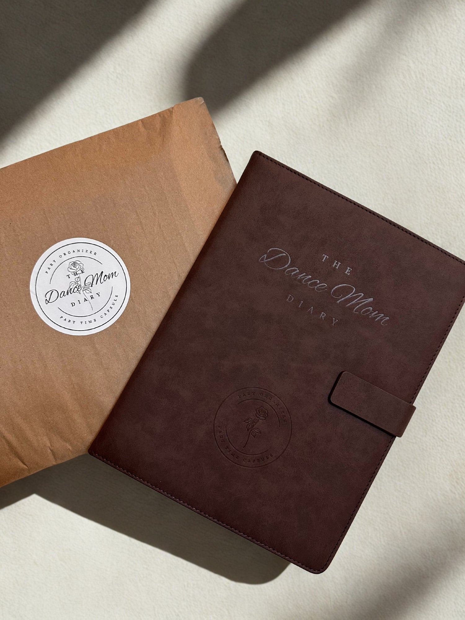 Brown leather Dance Mom Diary with embossed cover and strap closure beside branded kraft packaging behind a beige textured background with natural lighting.