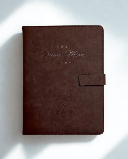 Brown leather Dance Mom Diary with embossed cover and strap closure on a soft white background with natural light shadows.